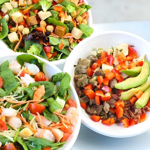 Three vibrant Bliss & Vinegar salads in white bowls, featuring fresh greens, proteins, and colorful vegetables.